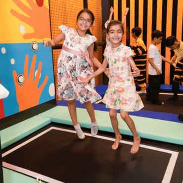 Two girls jumping together on an indoor trampoline at a kids play area.
